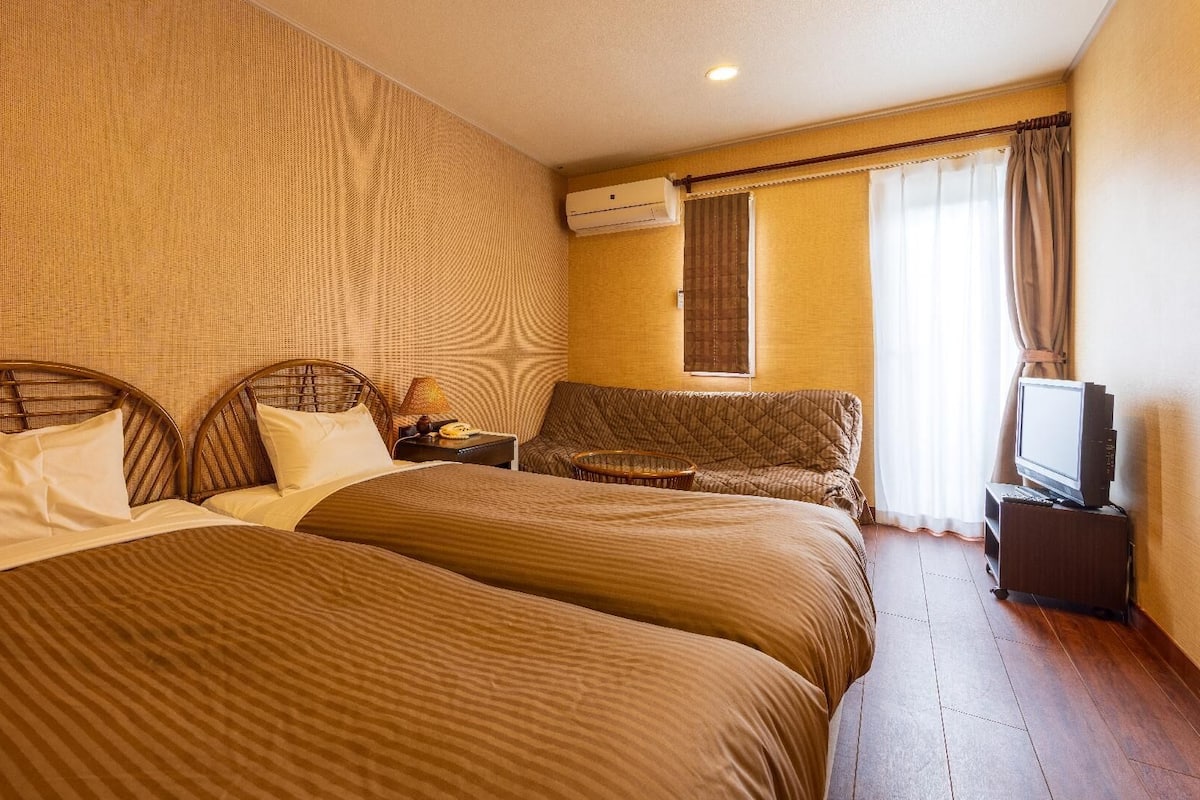 A hotel room with two beds, a sofa, a TV, and an air conditioning unit.
