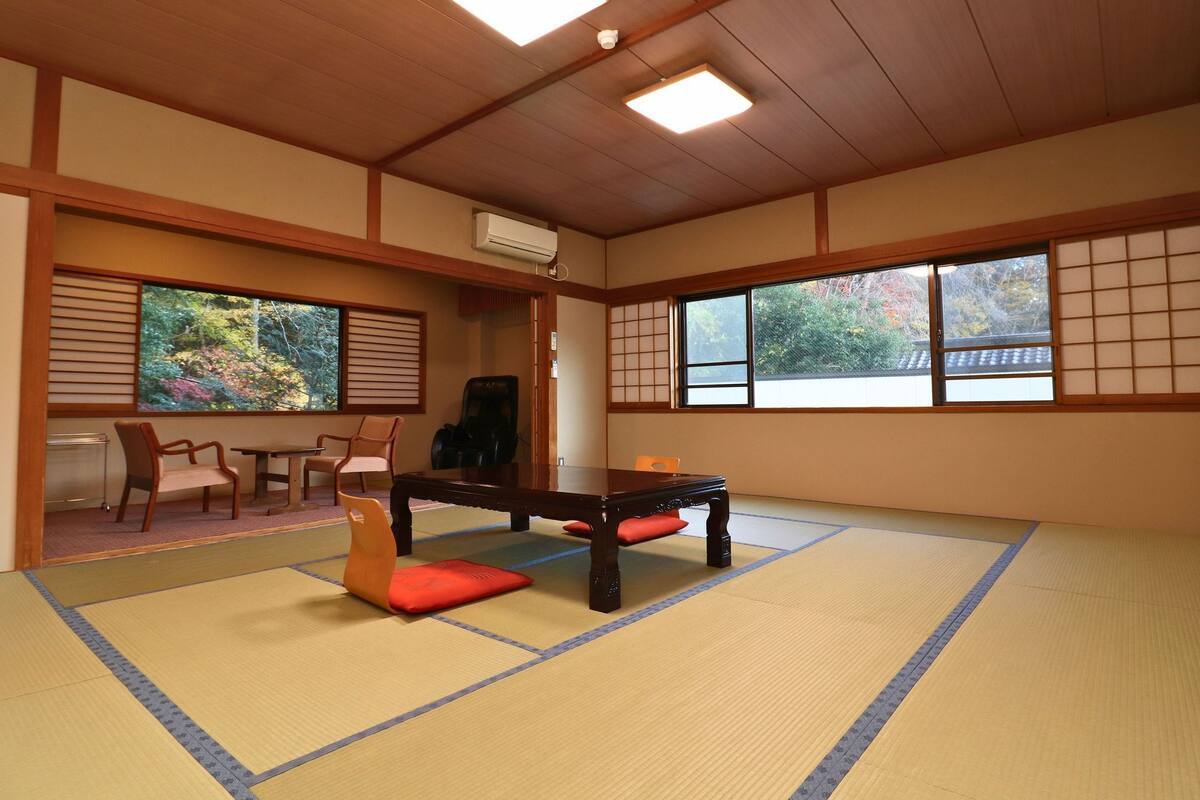 A tatami mat room with a tatami mat table, chairs, and a view of trees through large windows.