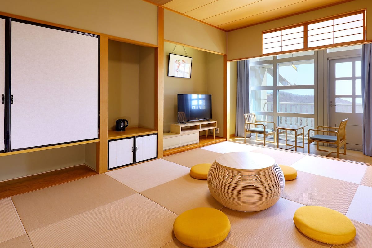 A traditional Japanese-style room with tatami flooring, a low table, yellow cushions, a television, and a sliding door with a view.