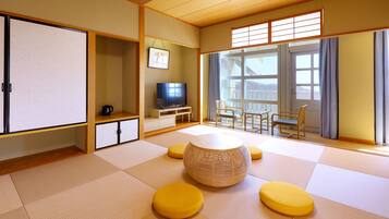 Una habitación tradicional japonesa con suelo de tatami, una mesa baja, cojines amarillos, un televisor y una puerta corredera con vistas.