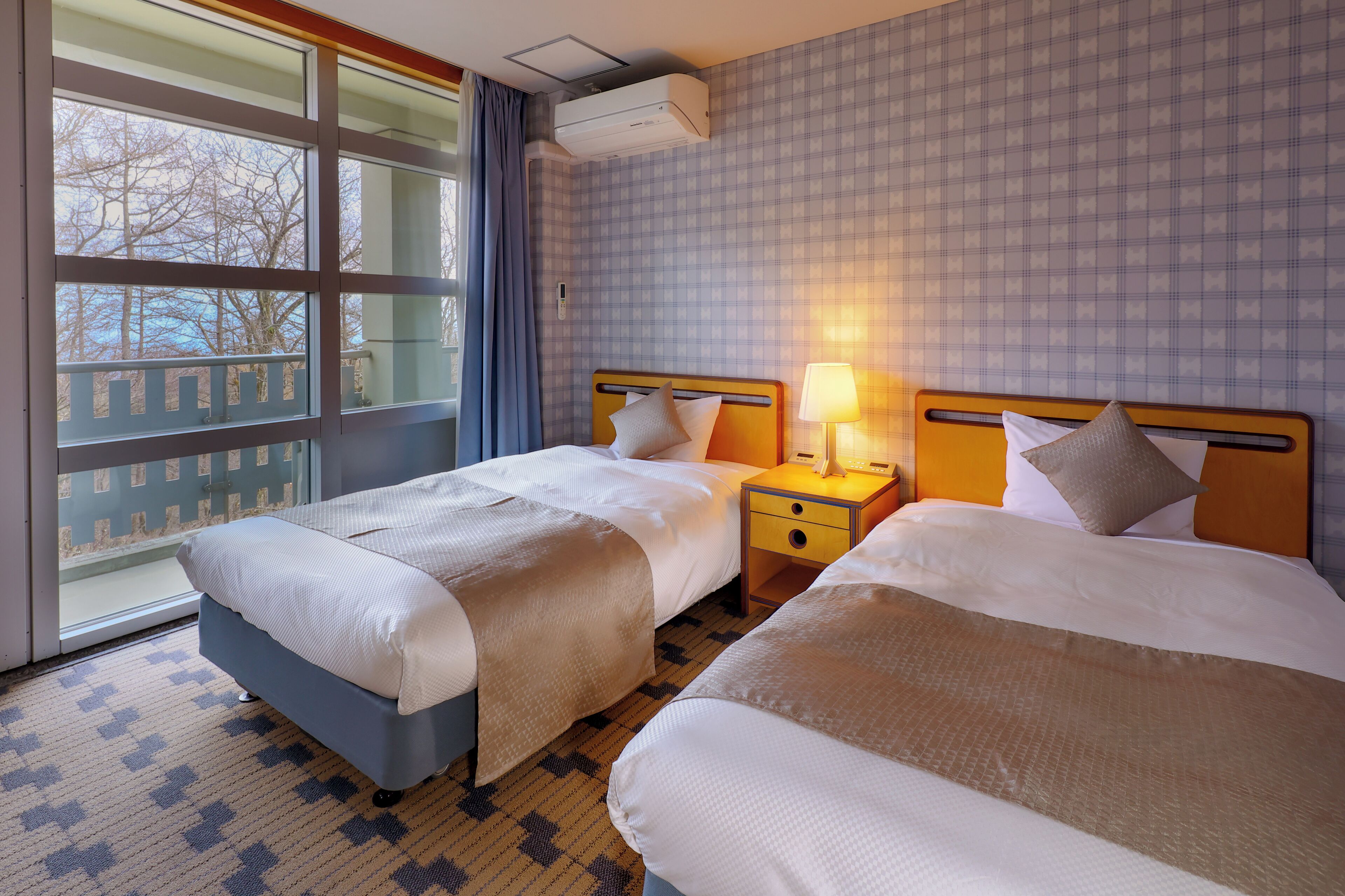 A hotel room with two beds, a window with a view, a bedside table with a lamp, and an air conditioning unit.