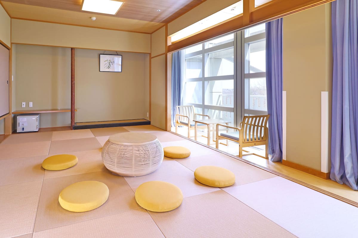 A tatami mat room with a central woven stool, four cushions, a small table, and chairs.