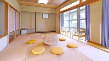 A tatami mat room with a central woven stool, four cushions, a small table, and chairs.