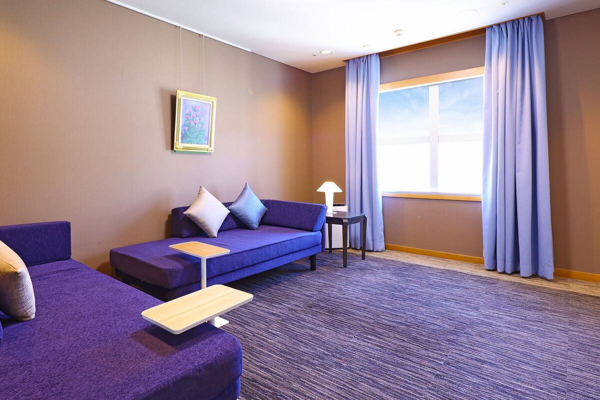 A hotel room with a purple sofa, a small table, and a window with curtains.