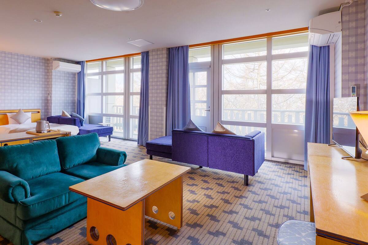 A hotel room with a teal sofa, a purple armchair, a wooden coffee table, and a desk with a lamp. There are large windows with curtains, an air conditioning unit, and a patterned carpet.