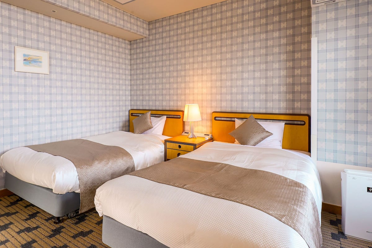 A hotel room with two beds, a nightstand with a lamp, a small refrigerator, and patterned wallpaper.