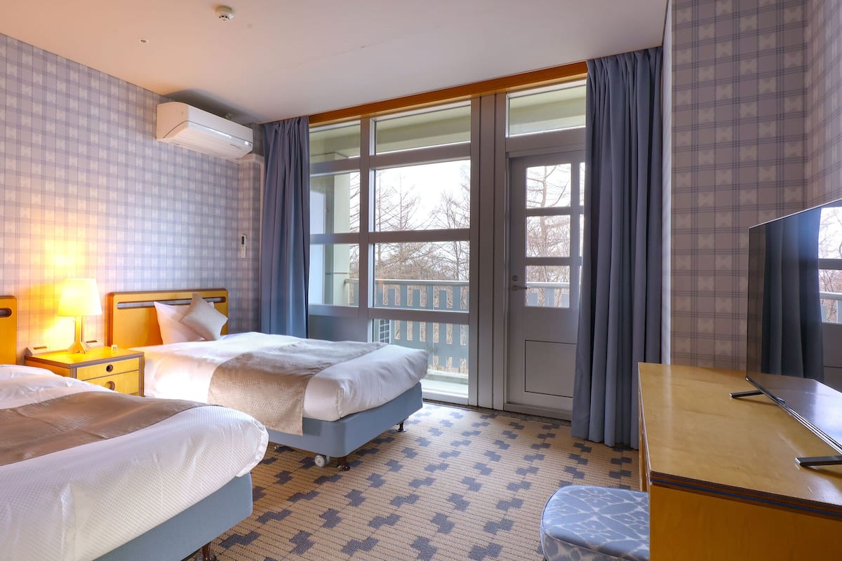 A hotel room with two beds, a desk, a TV, and a large window with curtains.