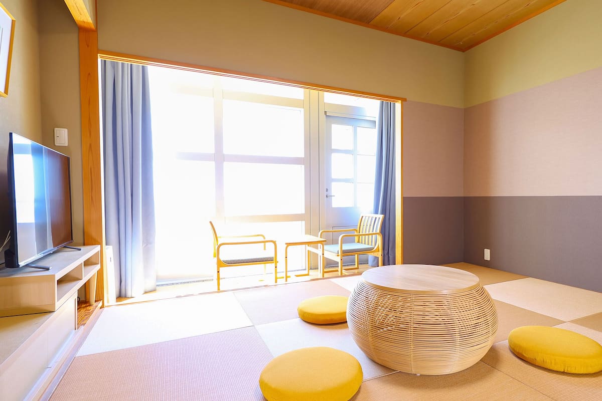 A room with tatami mats, a woven round table, yellow cushions, and a TV on a stand.
