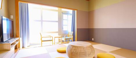 Una habitación con esteras de tatami, una mesa redonda tejida, cojines amarillos y un televisor sobre un mueble.