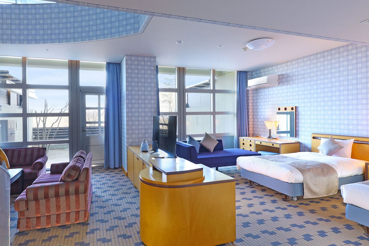 A hotel room with a bed, a sofa, a desk, and a chair. There are large windows and an air conditioning unit on the wall.
