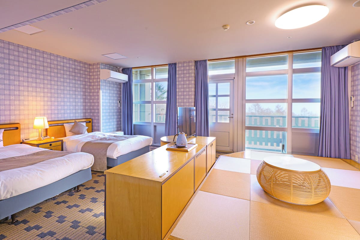 A hotel room with two beds, a desk, a lamp, and a view of the outside.