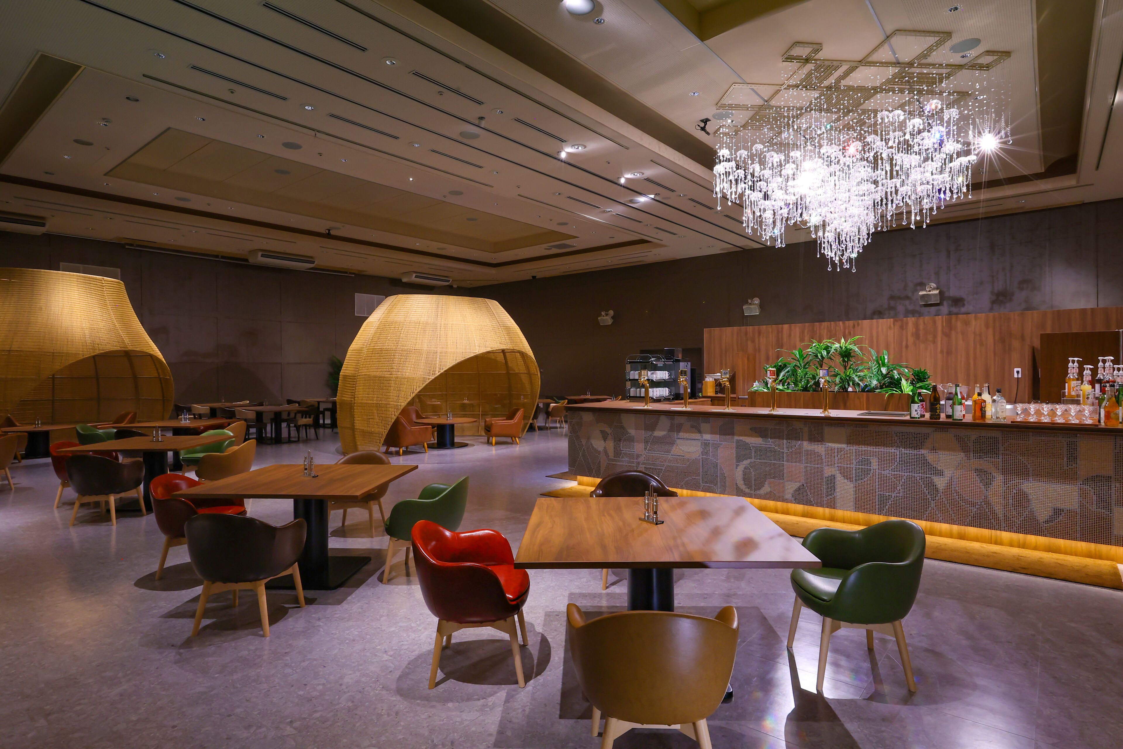 A modern restaurant with wooden tables and chairs, a bar area with a green plant, and a chandelier.