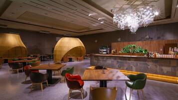 A modern restaurant with wooden tables and chairs, a bar area with a green plant, and a chandelier.