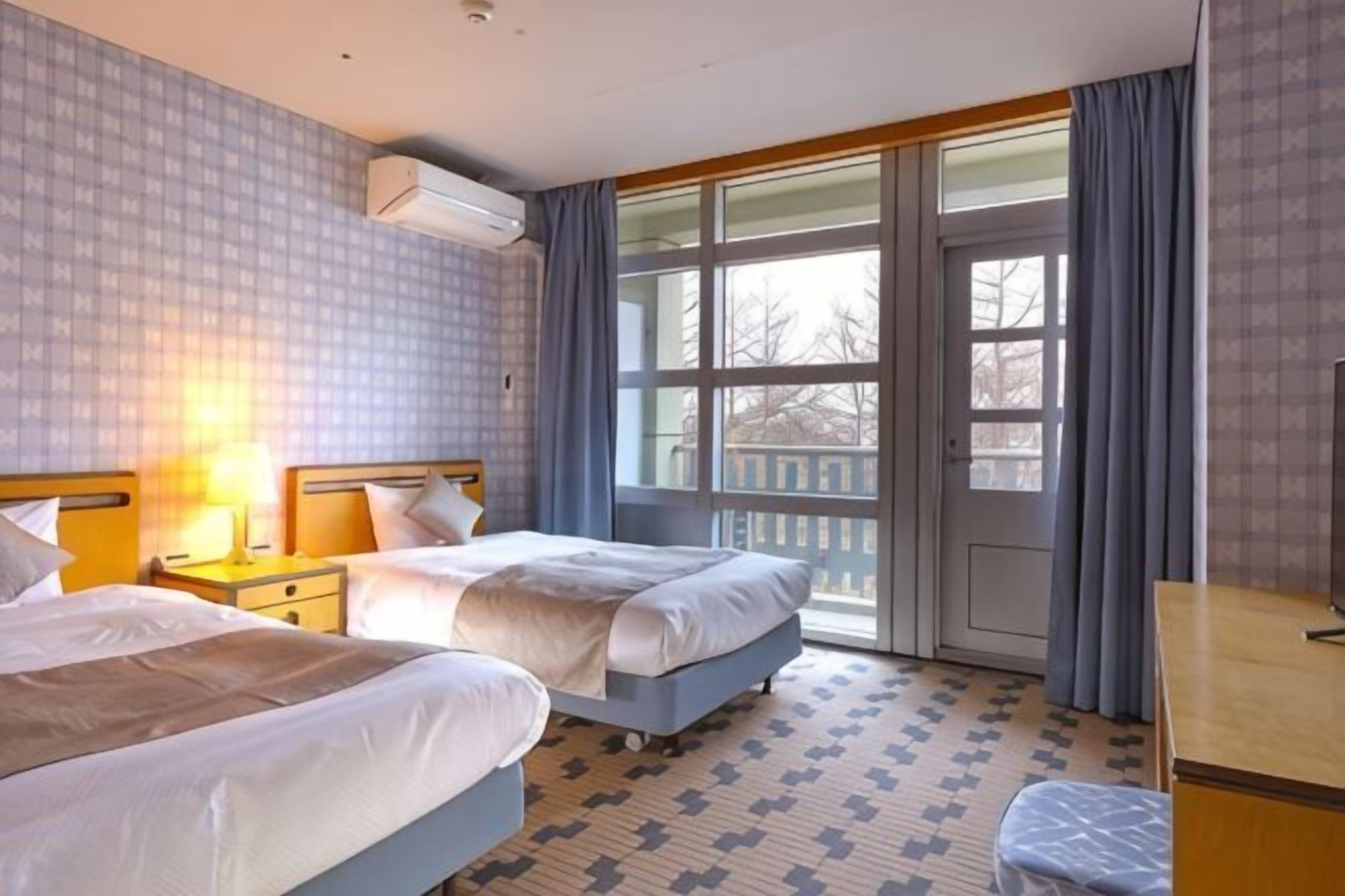 A hotel room with two beds, a desk, a chair, and a large window with curtains.