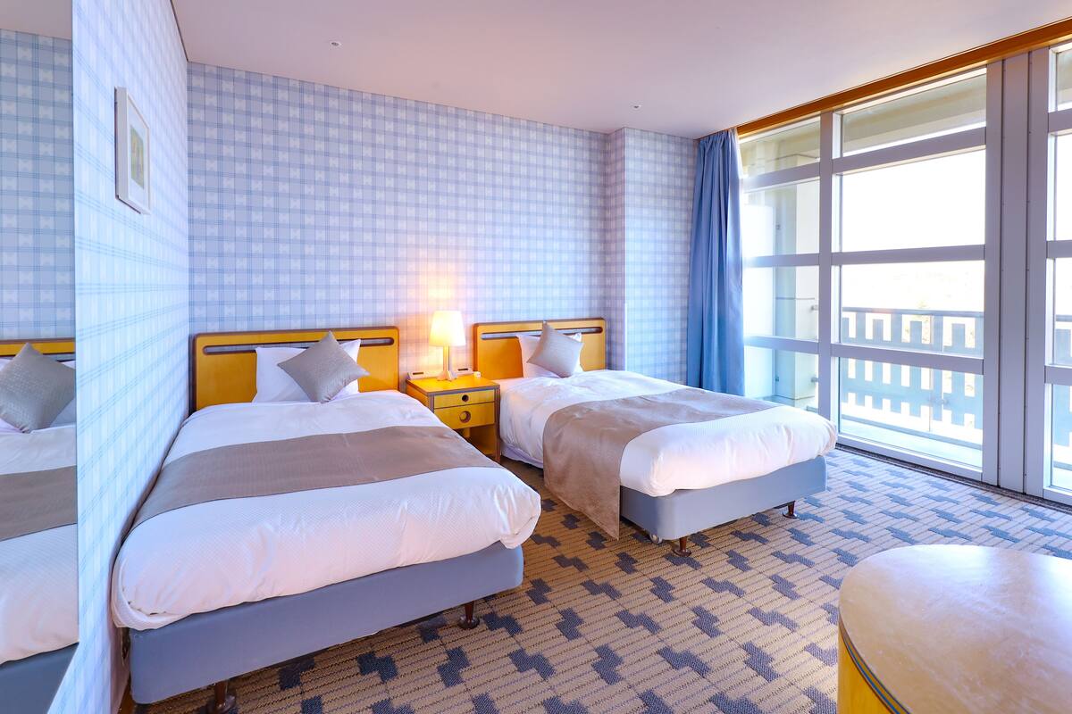 A hotel room with two beds, a small table, and a large window with a view.