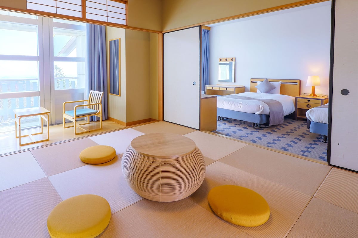A hotel room with tatami flooring, a bed, a bedside table, a lamp, a chair, and a view through a sliding door.