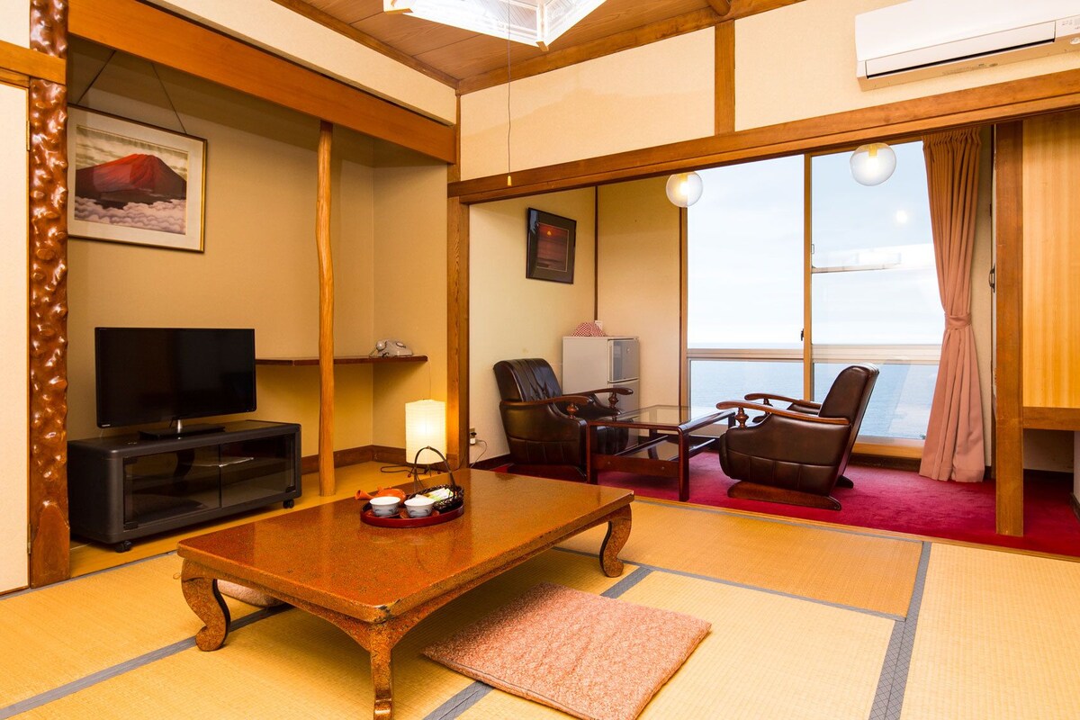Una habitación tradicional japonesa con suelo de tatami, televisor, escritorio con lámpara y vistas al mar a través de amplias ventanas.