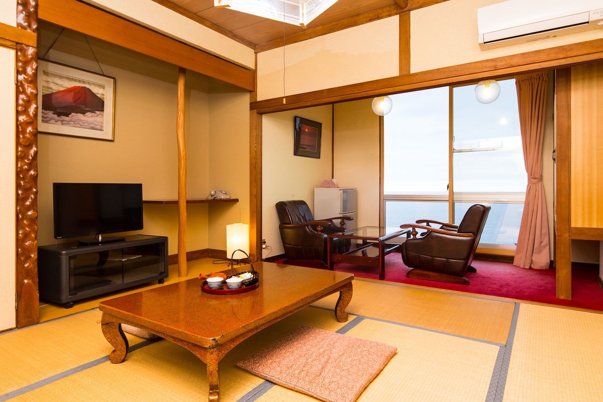 Een traditionele Japanse kamer met tatami vloeren, een televisie, een bureau met lamp en uitzicht door een schuifdeur.