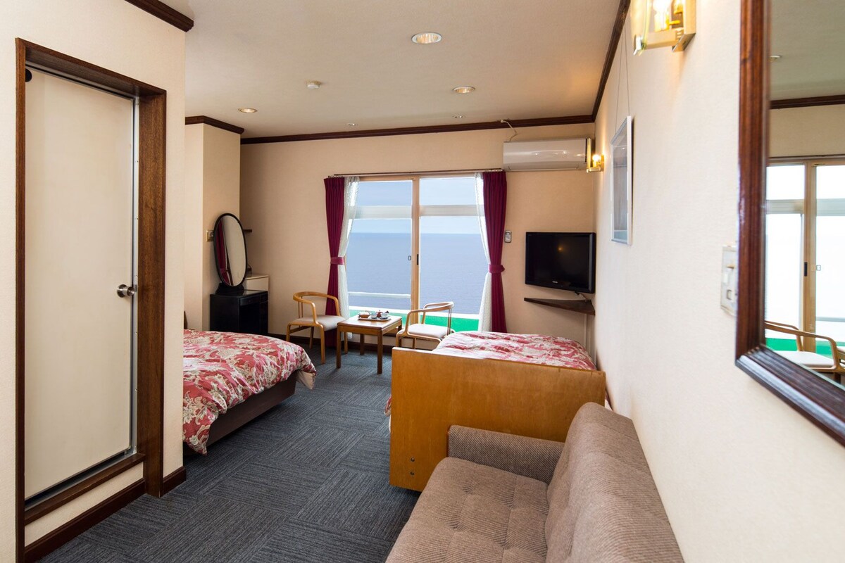 Habitación de hotel con dos camas, un escritorio, un televisor y una ventana con vistas al mar.