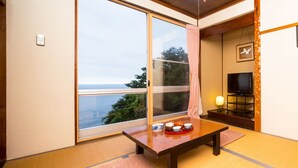A room with a large sliding glass door leading to a balcony with a view of the sea. There is a small table with a tray of tea and cups, a television on a stand, and a framed picture on the wall.