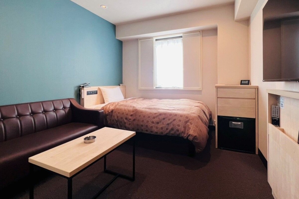 A compact hotel room with a bed, a small table, a sofa, a TV, and a mini-fridge.
