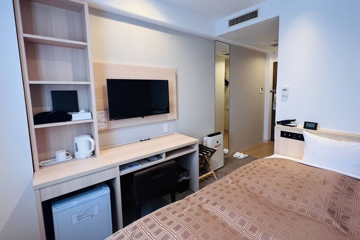 A hotel room with a bed, a desk with a TV, a coffee maker, a small fridge, and a wardrobe.