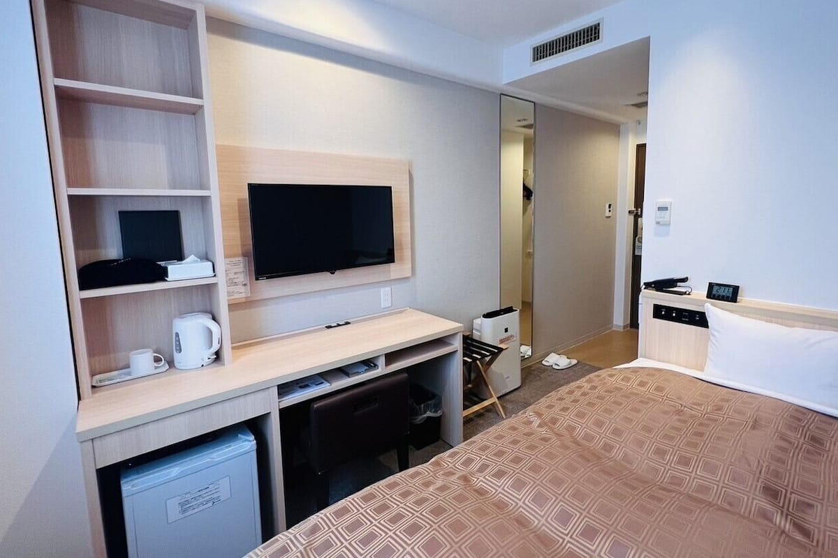 A hotel room with a bed, a desk with a TV, a coffee maker, a small fridge, and a wardrobe.