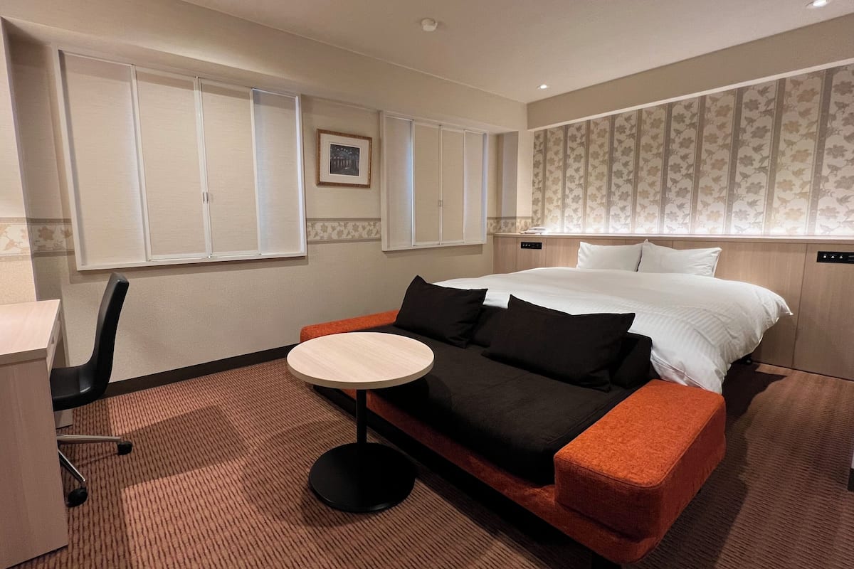 A hotel room with a bed, a small round table, a sofa, a chair, and a wardrobe.