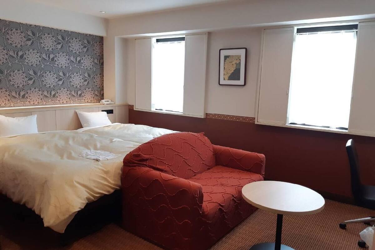 A hotel room with a bed, a red armchair, a round table, and a window with blinds.