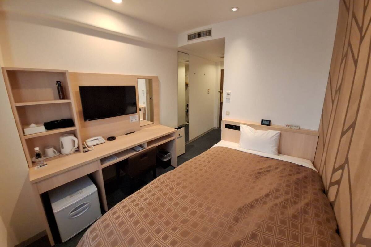 A hotel room with a bed, a desk with a computer, a TV, and a small kitchenette.