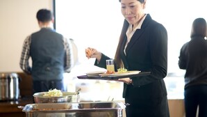 Una persona serve del cibo in una stanza con una finestra, un tavolo per buffet e un uomo sullo sfondo.