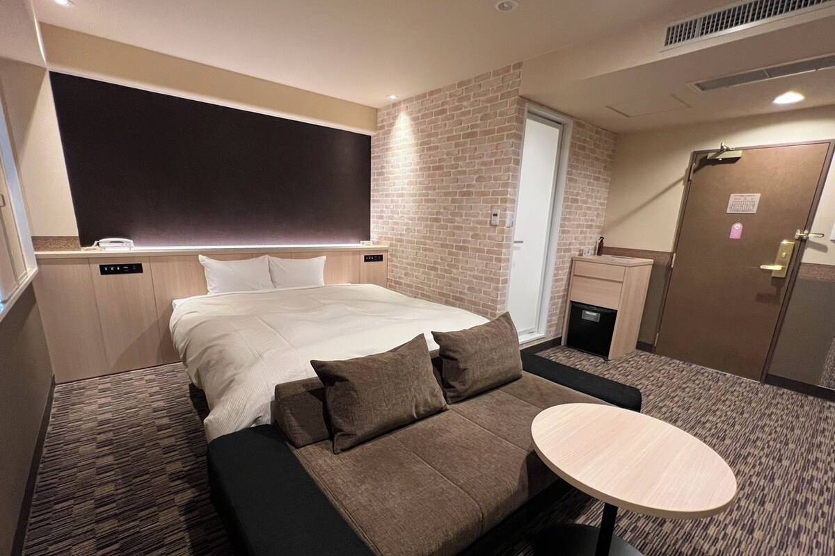 A hotel room with a bed, a sofa, a small table, a brick wall, a door, and a window.