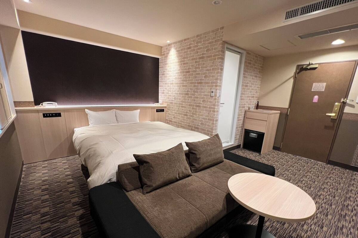 A hotel room with a bed, a sofa, a small table, a brick wall, a door, and a window.