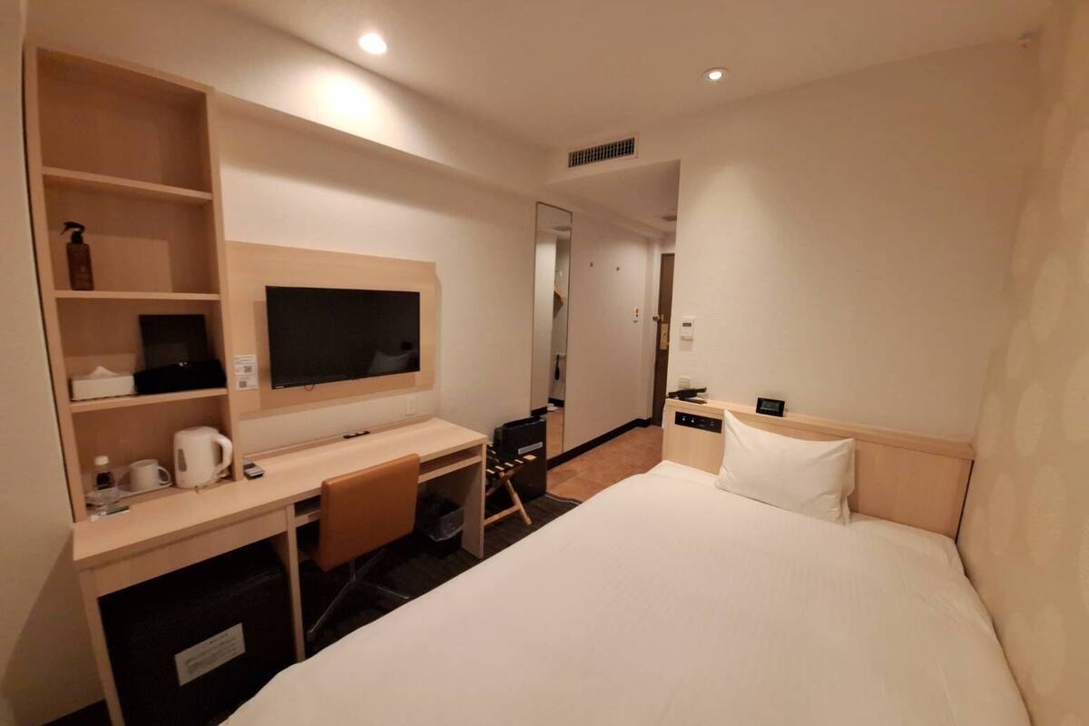 A hotel room with a bed, a desk with a television, a mirror, and a small bathroom area.
