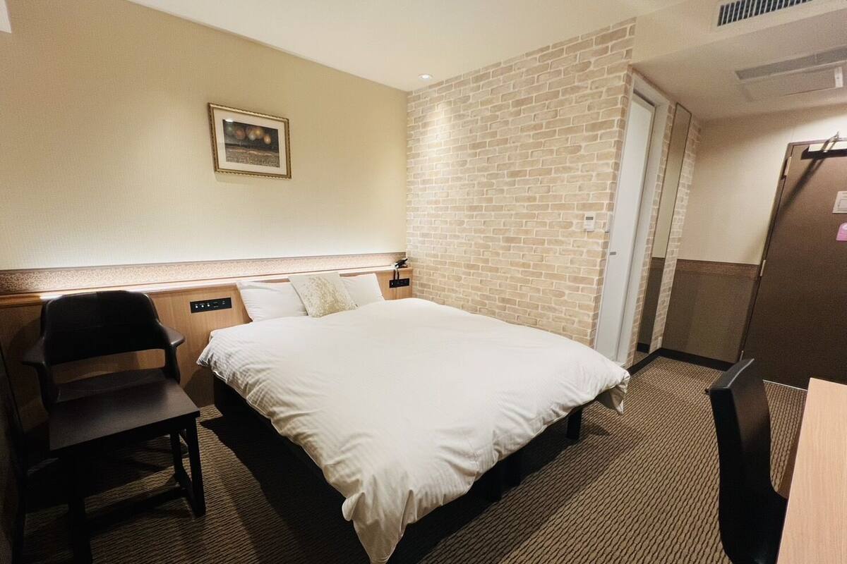 A hotel room with a bed, a desk, a chair, a picture on the wall, and a brick-patterned wall.