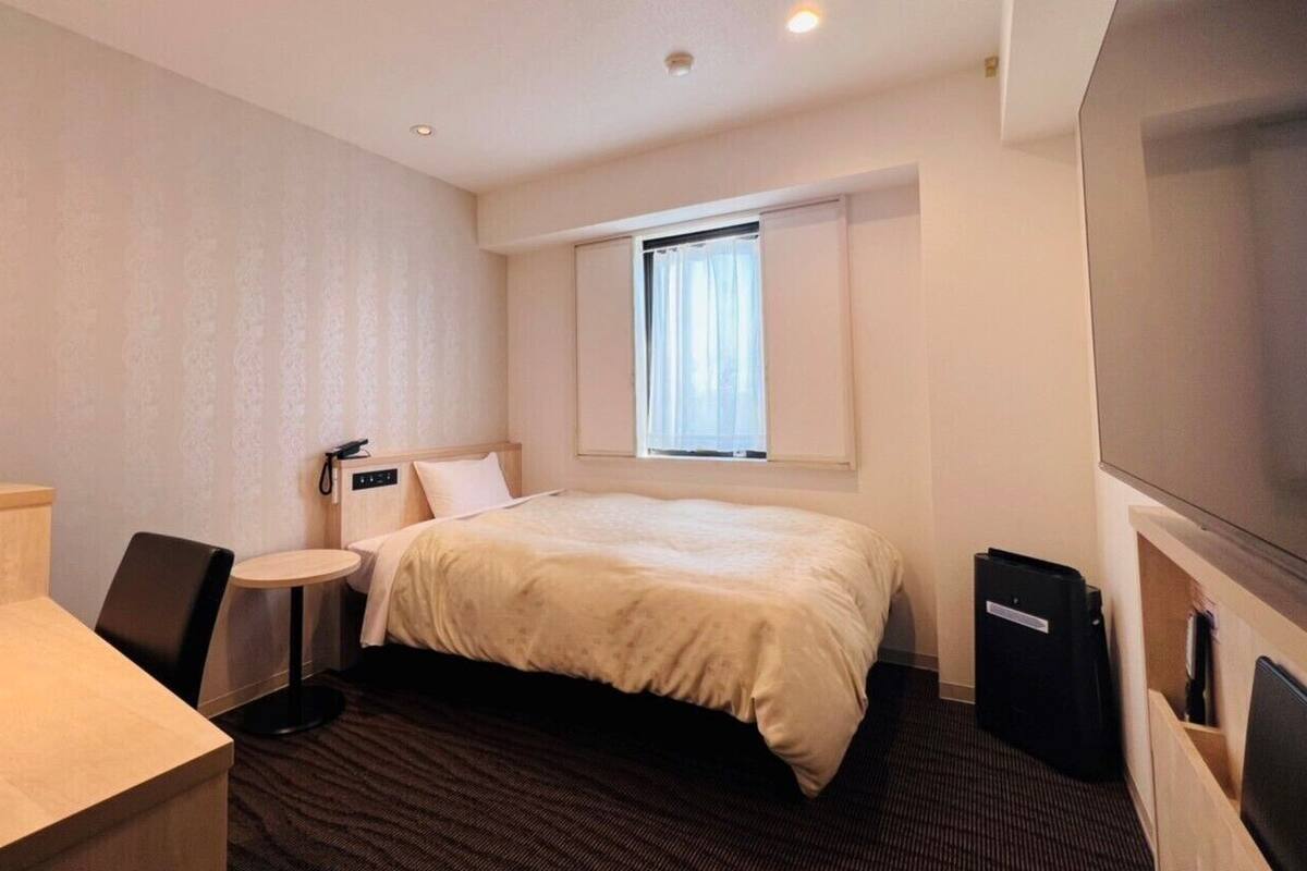 A hotel room with a bed, a desk, a chair, a small round table, a TV, and a window with curtains.