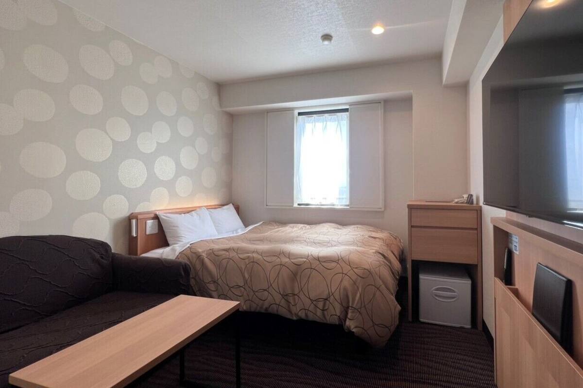 A compact hotel room with a bed, a small desk, a TV, and a window with curtains.