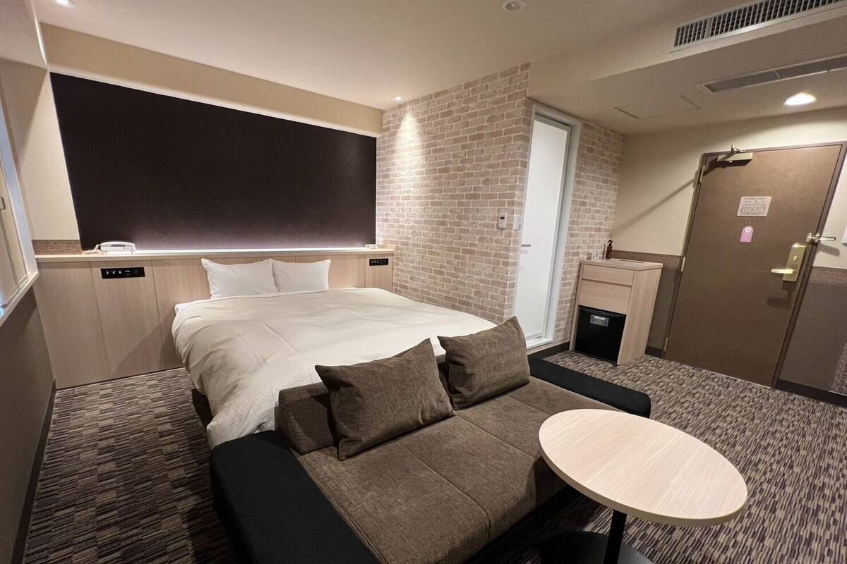A hotel room with a bed, a sofa, a small table, a brick wall, a door, and a window.