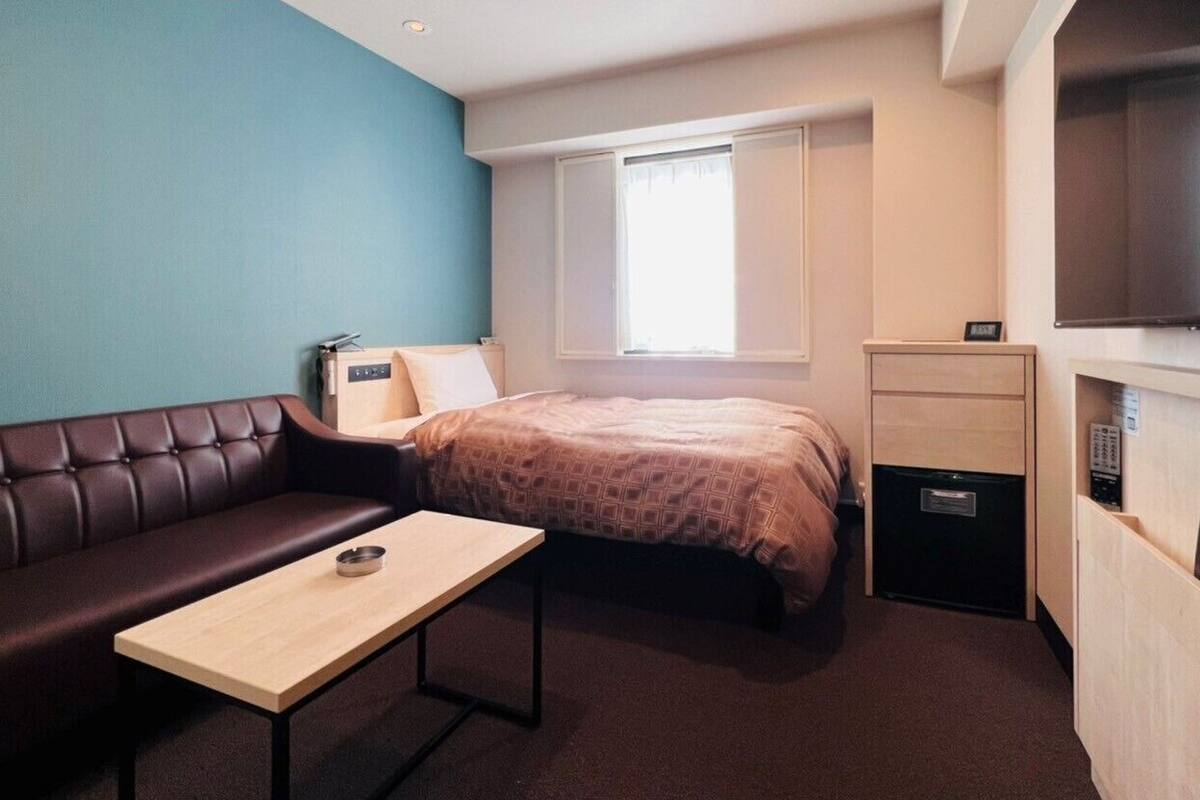 A compact hotel room with a bed, a small table, a sofa, a TV, and a mini-fridge.