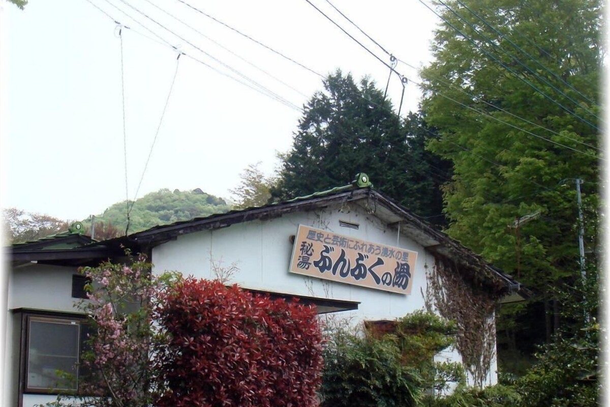 A small building with a sign in Japanese, surrounded by greenery and a hill in the background.