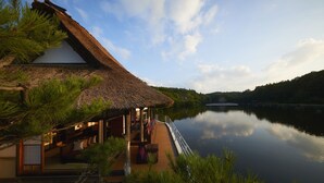 A thatched-roof house with a wooden deck overlooking a calm lake, surrounded by greenery. - Hoshino Resorts Aomoriya (Rokunohe)