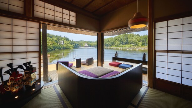 A traditional Japanese-style room with tatami flooring, a low platform bed with colorful cushions, and a view of a lake and forest. - Hoshino Resorts Aomoriya (Rokunohe)