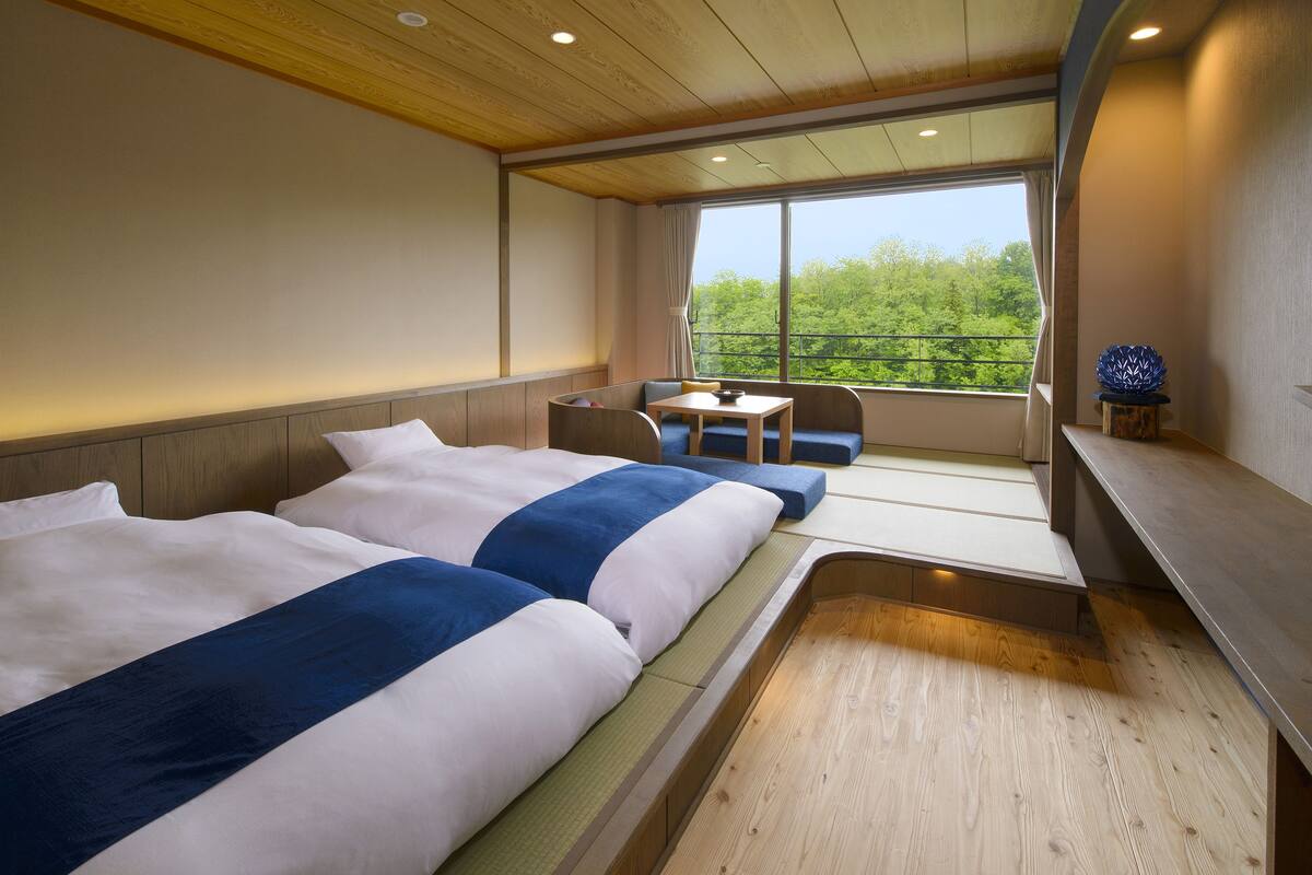 A hotel room with two beds, a small table, and a view of greenery through the window.
