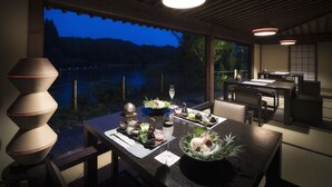 A dining area with a view of a lake at dusk, featuring a wooden table set with traditional Japanese dishes, a glass of sake, and a decorative vase. - Hoshino Resorts Aomoriya (Rokunohe)