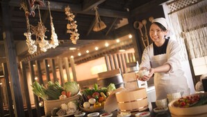 A kitchen with hanging light fixtures, a person in a chef's uniform, and various food items and cooking utensils on the counter. - Hoshino Resorts Aomoriya (Rokunohe)