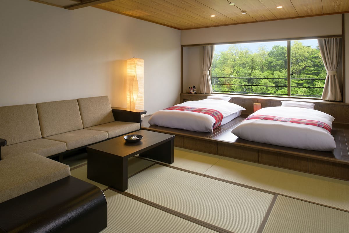 A modern hotel room with a sofa, two beds with white bedding, a bedside table, a lamp, and a view of trees through the window.