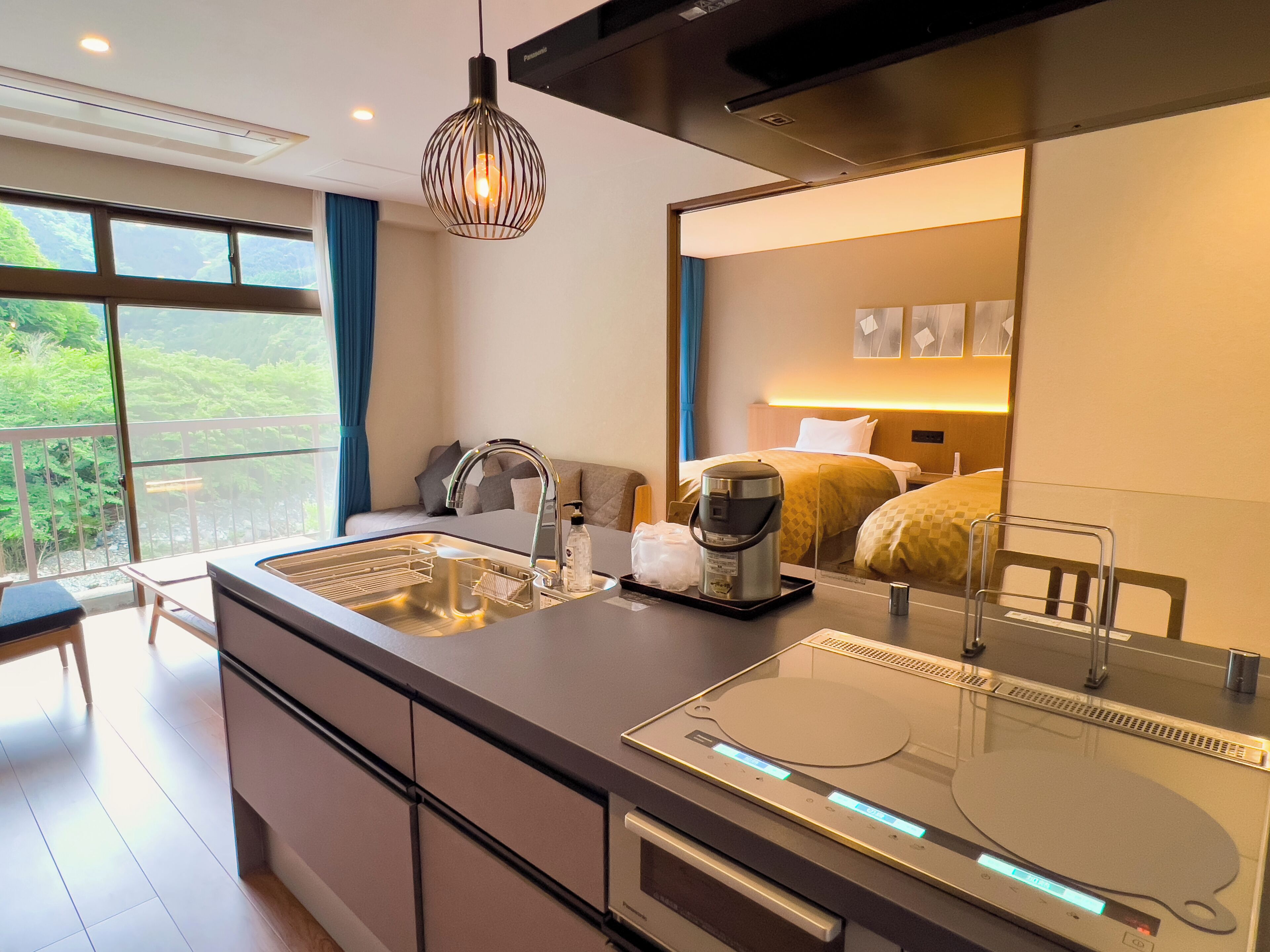 Une chambre d’hôtel moderne dotée d’un coin cuisine, d’un lit avec une tête de lit et offrant une vue sur la verdure environnante.