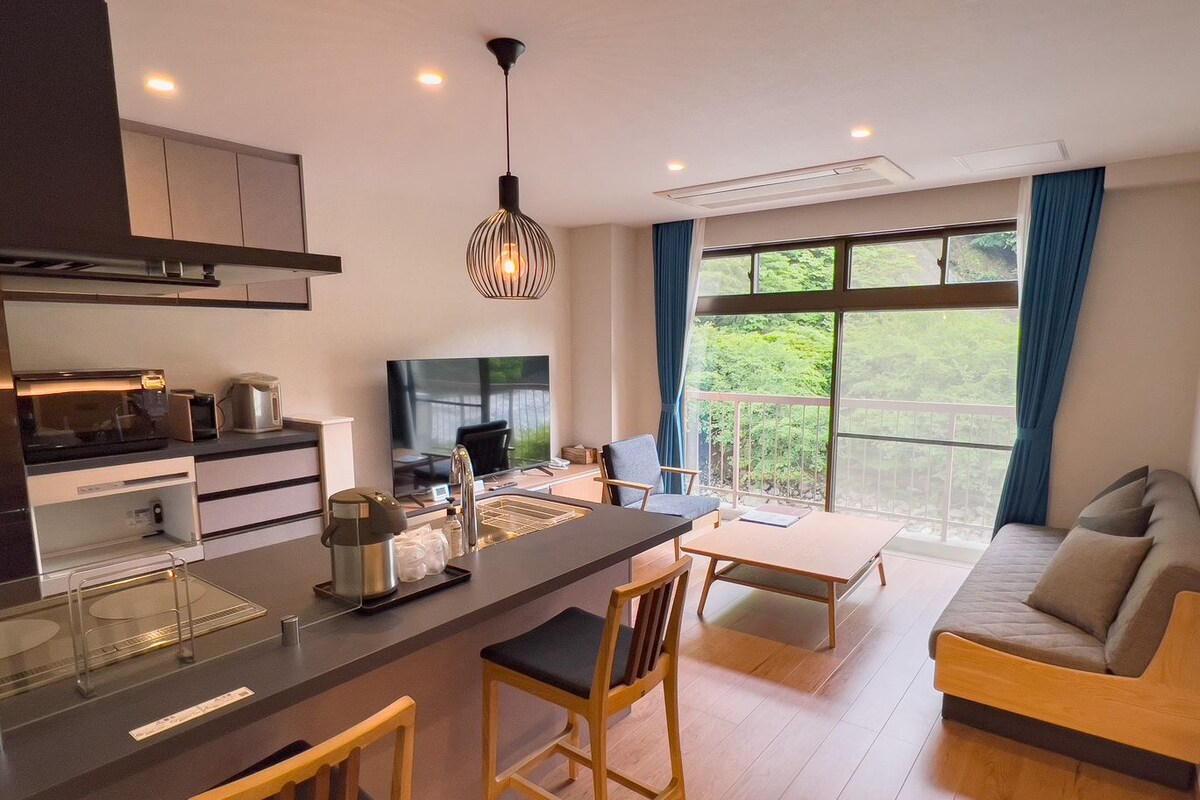Une cuisine moderne dotée d’un îlot central, un coin repas avec une table et des chaises, un canapé et une grande fenêtre donnant sur un paysage verdoyant.