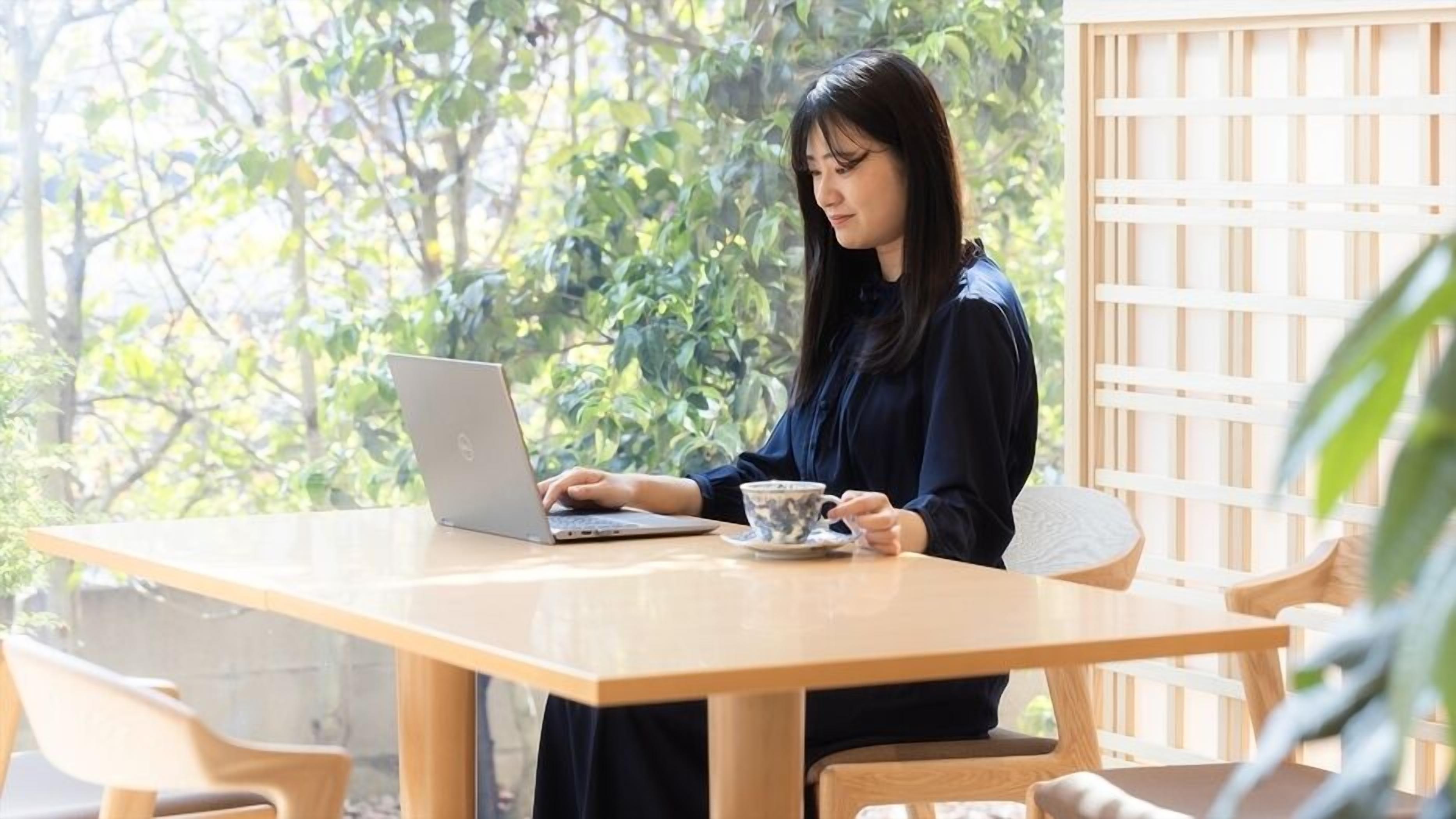 A person using a laptop at a wooden table with a cup, in a room with a wooden lattice wall and greenery outside.