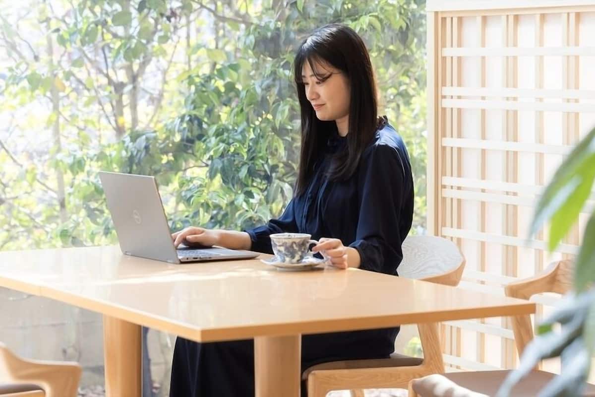 A person using a laptop at a wooden table with a cup, in a room with a wooden lattice wall and greenery outside.